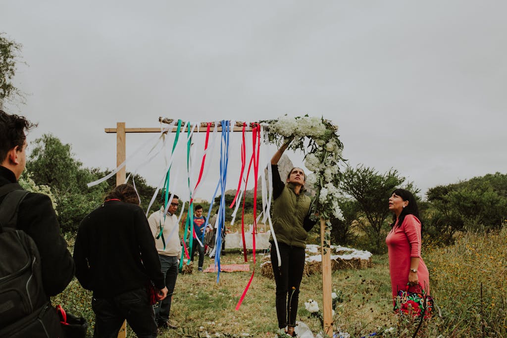 People preparing a colorful outdoor wedding arch with ribbons and flowers in a natural setting.